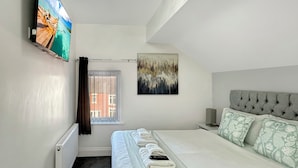 A modern hotel room with a large bed, a flat-screen TV mounted on the wall, a ceiling light fixture, and a window with a view of buildings outside. - Granada Apartments Holmfield Rd flat 4 2nd floor loft type (Blackpool)