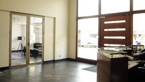 A reception area with a glass desk, a large window with a view of the street, and a sliding glass door leading to a room with a tripod lamp. - Living Pastene  (Santiago)