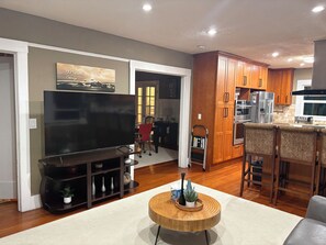 Una sala de estar con televisor de pantalla plana, una mesa de centro de madera y una cocina con armarios de madera y refrigerador.
