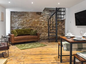 Una sala acogedora con una pared de piedra, un sofá de cuero marrón, un plaid verde, una mesa de comedor de madera dispuesta con platos y vasos blancos, y una escalera de caracol.