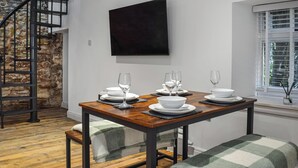 Un comedor con una mesa de madera dispuesta para cuatro, pared de piedra, escalera y ventana con persianas.