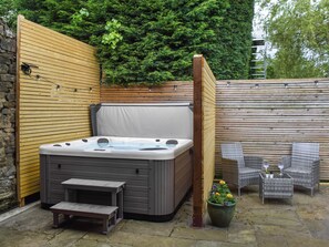 A hot tub in a backyard with a wooden fence, a stone wall, and wicker furniture.