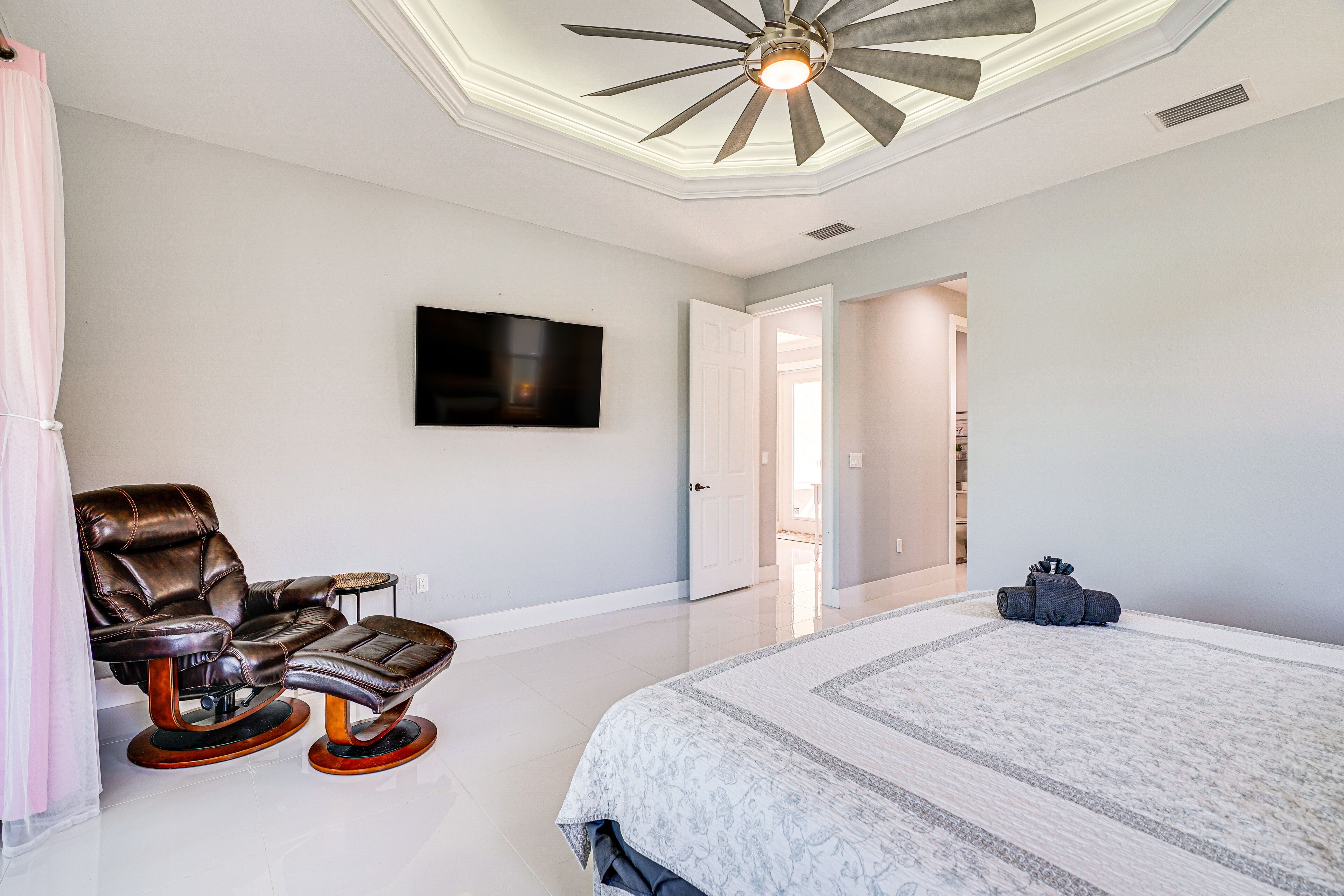 A bedroom with a large bed, a reclining chair, a flat-screen TV, and a ceiling fan.