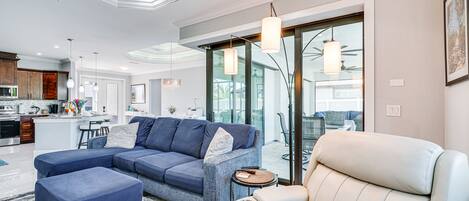 Una sala moderna con un sofá seccional, un sillón reclinable y una mesa de centro. Se observa una cocina al fondo con una isla grande y taburetes de bar.