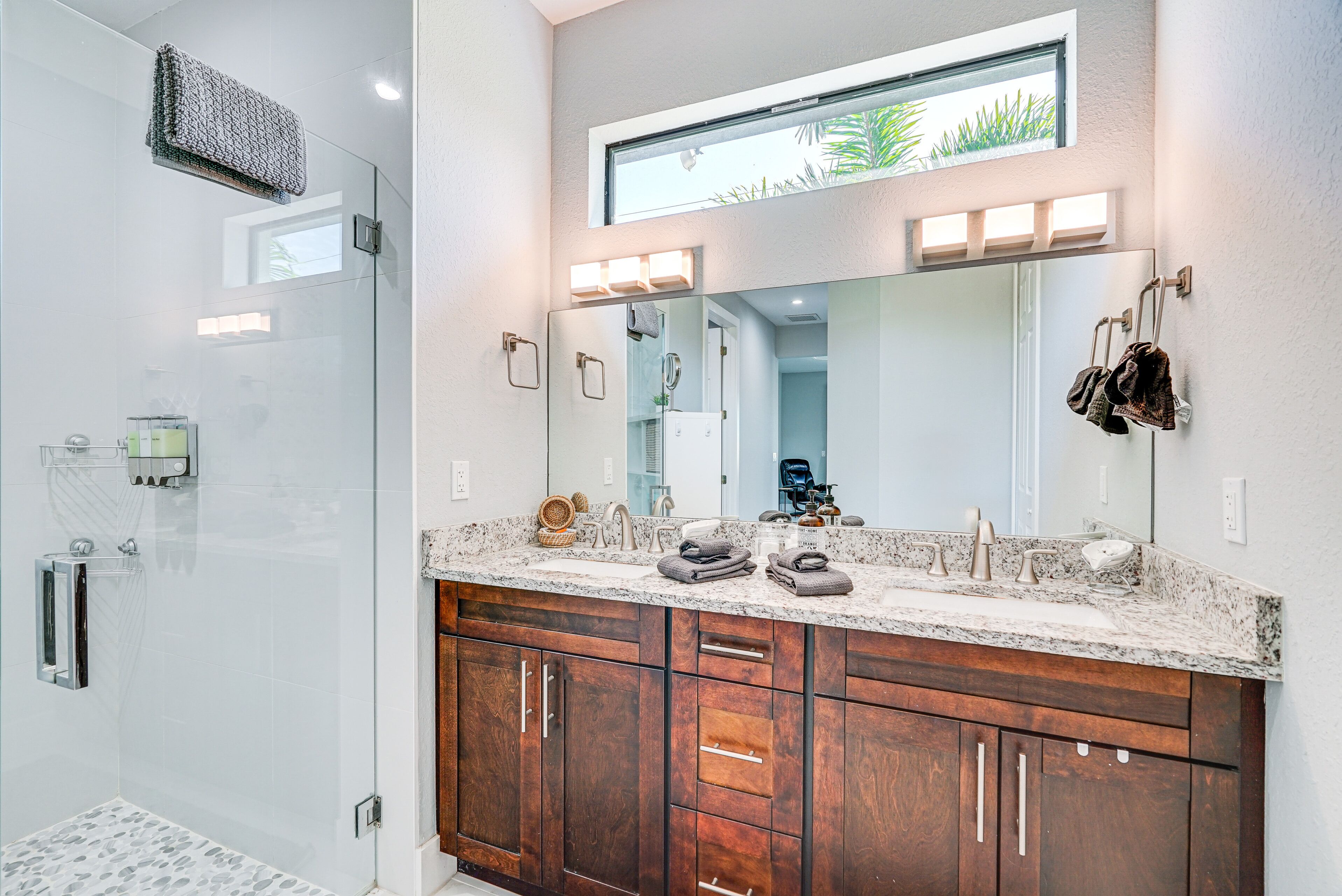 Un baño moderno con cabina de ducha de vidrio, un mueble de tocador espacioso con encimera de granito y un espejo grande sobre el lavabo.