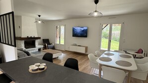 Una cocina moderna con una isla central, comedor con sillas blancas, televisor montado en la pared y chimenea.