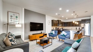 Ein modernes Wohnzimmer mit einem grauen Sofa, einem Couchtisch und einem Fernseher. Es gibt eine Küchenzeile mit dunklen Schränken und einem Edelstahlkühlschrank.
