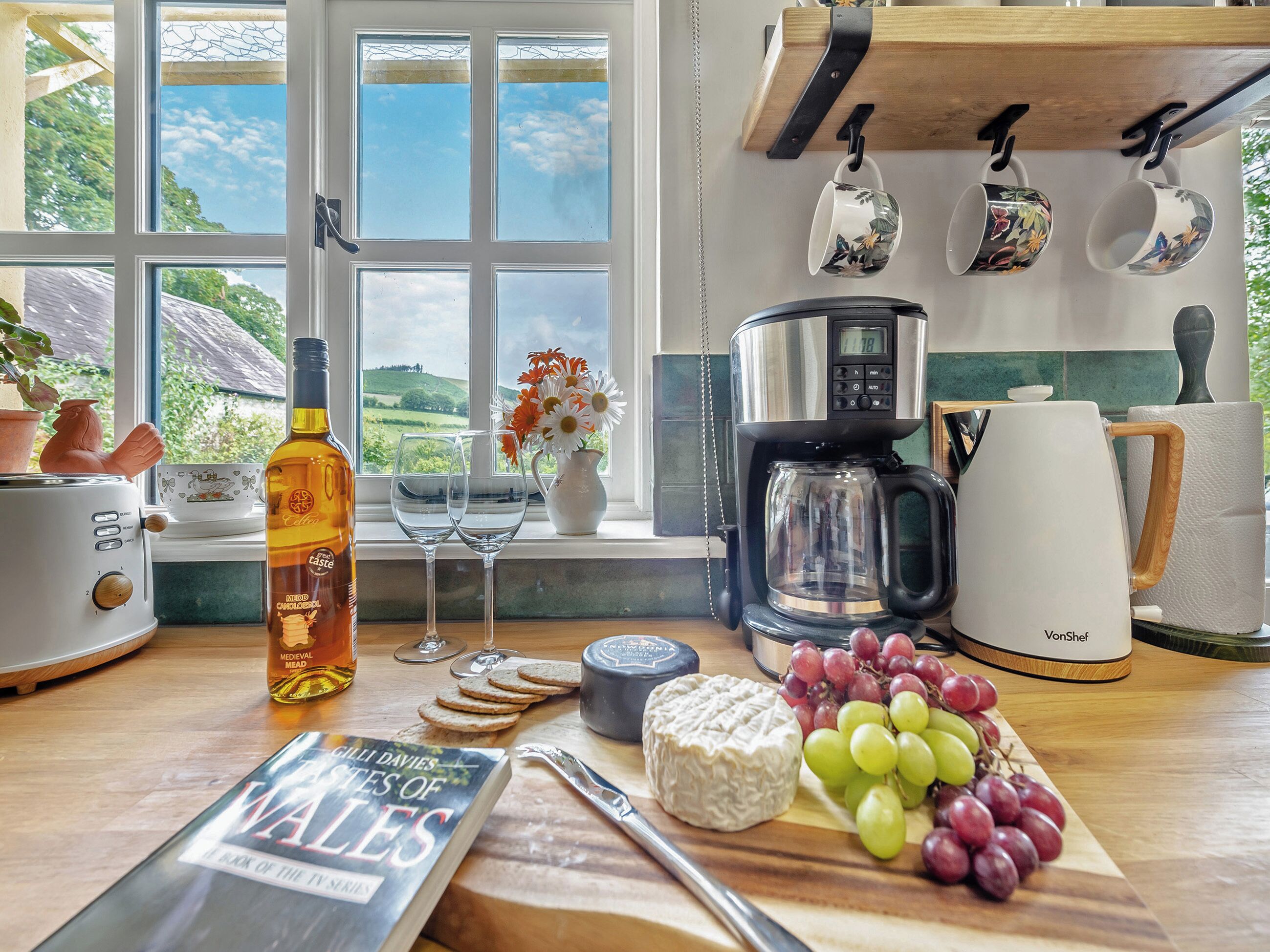 Une cuisine équipée d’une machine à café, d’une bouilloire, d’un livre, d’une bouteille et d’une fenêtre donnant sur la campagne.