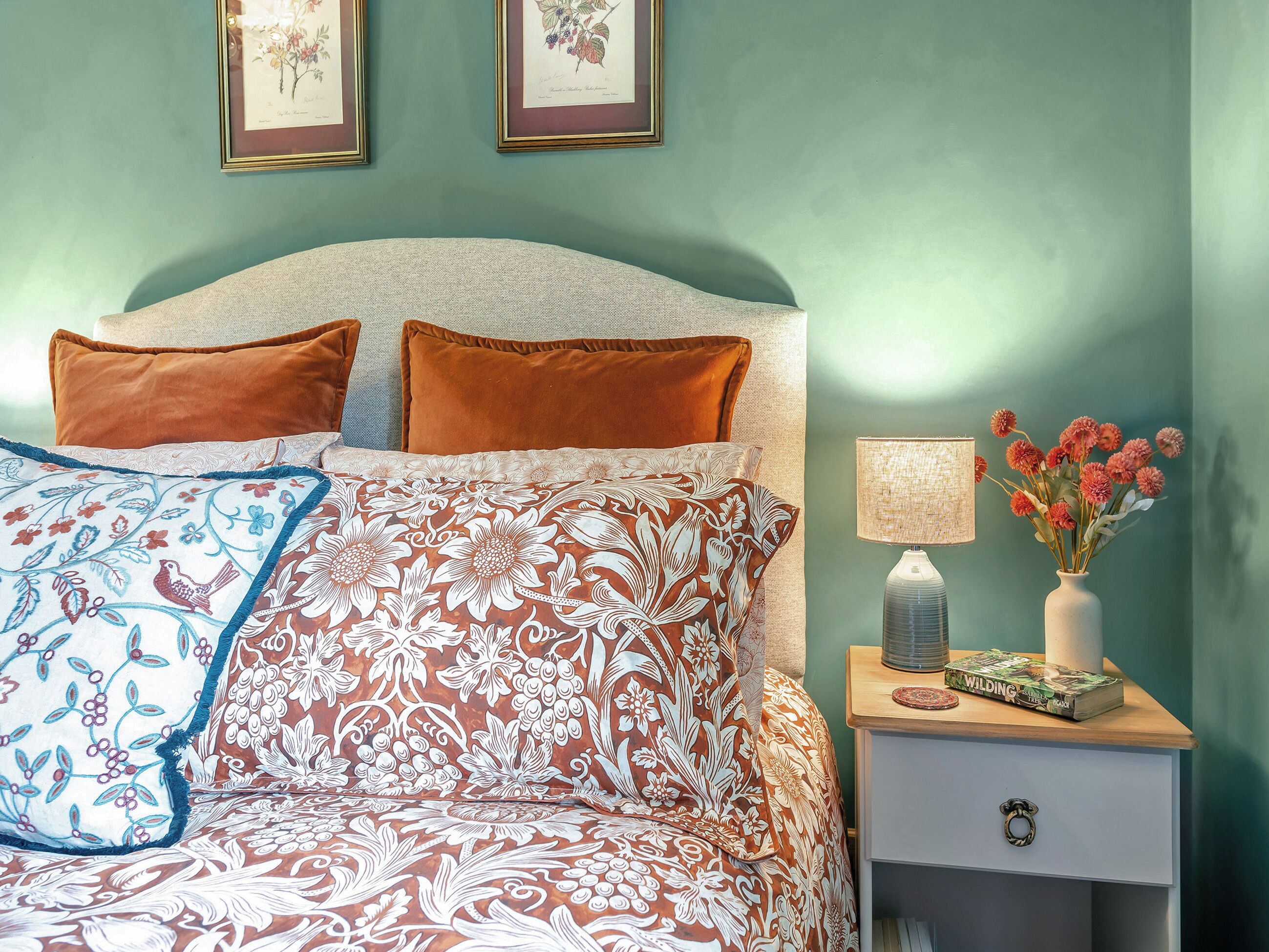 A neatly made bed with patterned bedding, a bedside table with a lamp and a vase of flowers, and two framed botanical prints on the wall.