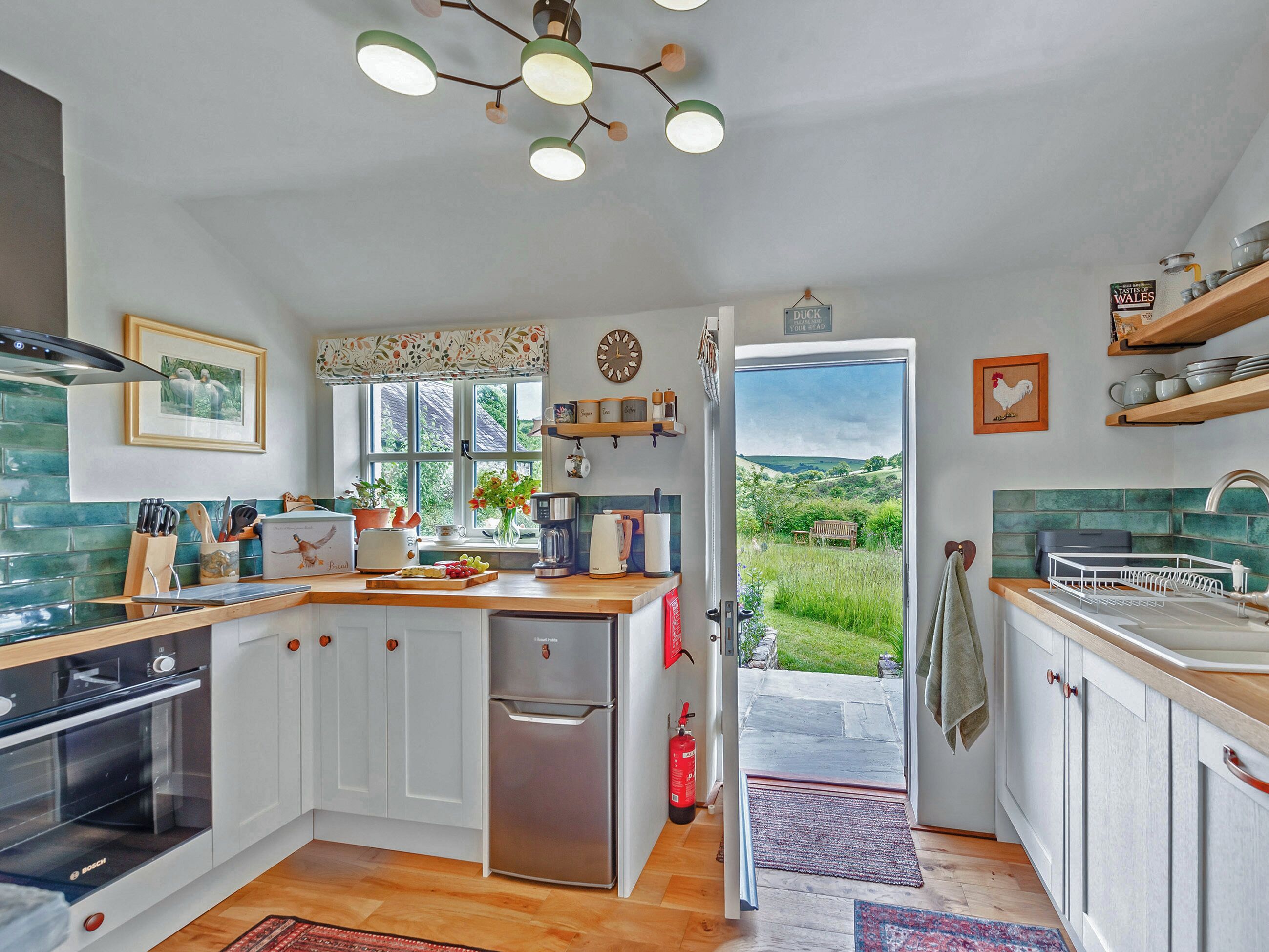A kitchen with white cabinets, a Bosch oven, a coffee machine, and a view through an open door to a garden.