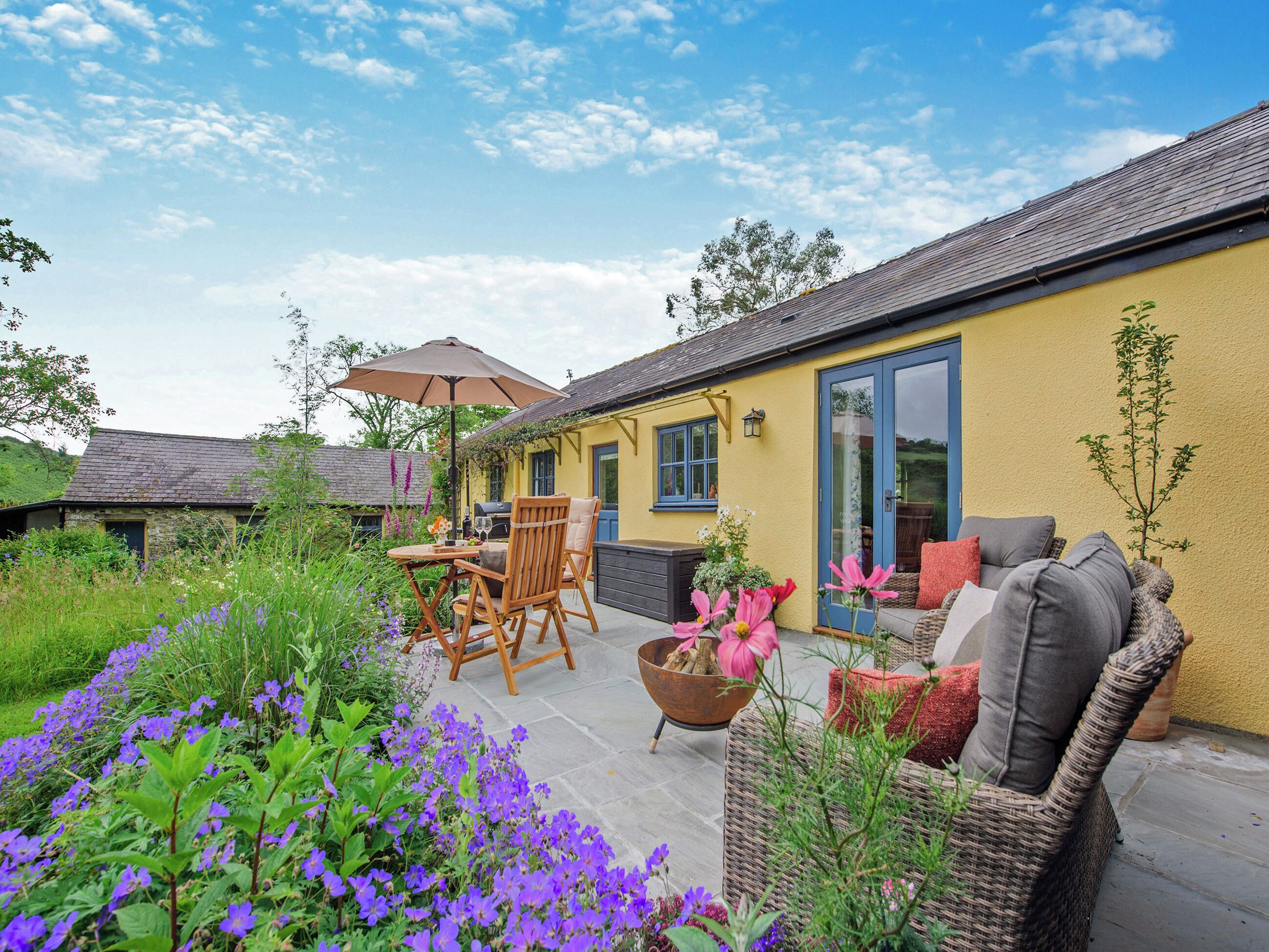 Una casa gialla con un patio, mobili da esterno e un giardino con fiori viola.
