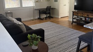 Ett vardagsrum med en svart soffa, ett soffbord med en växt, en tv på ett träställ och en mönstrad matta.