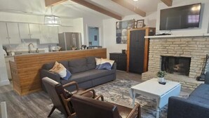 Una sala moderna con sofá, mesa de centro y sillones. Hay una chimenea, televisión y una zona de cocina al fondo.