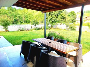 An outdoor patio with a wooden table and wicker chairs, a covered area, and a view of a well-maintained backyard with a white fence and greenery.