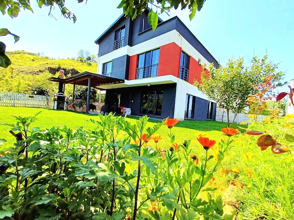 A modern two-story house with a covered patio, surrounded by a well-maintained garden with various plants and flowers.