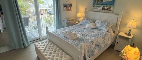 A bedroom with a bed, a bench, a nightstand, a lamp, a painting, and a view of the outside.