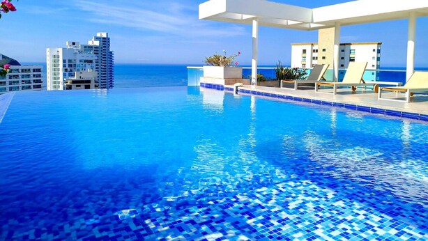 Une piscine d’un bleu éclatant, bordée de carreaux, avec des chaises longues et une vue sur l’océan et les bâtiments qui se dressent à l’arrière-plan.