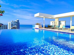 A clear blue pool with a tiled edge, lounge chairs, and a view of the ocean and buildings in the background.