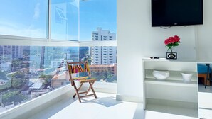 Un salon moderne doté d’une grande fenêtre, d’un téléviseur à écran plat, d’une chaise en bois et d’une petite étagère blanche ornée d’objets décoratifs.