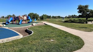 Un parque infantil con toboganes y estructuras para escalar, rodeado de un césped cuidado y un cielo despejado.