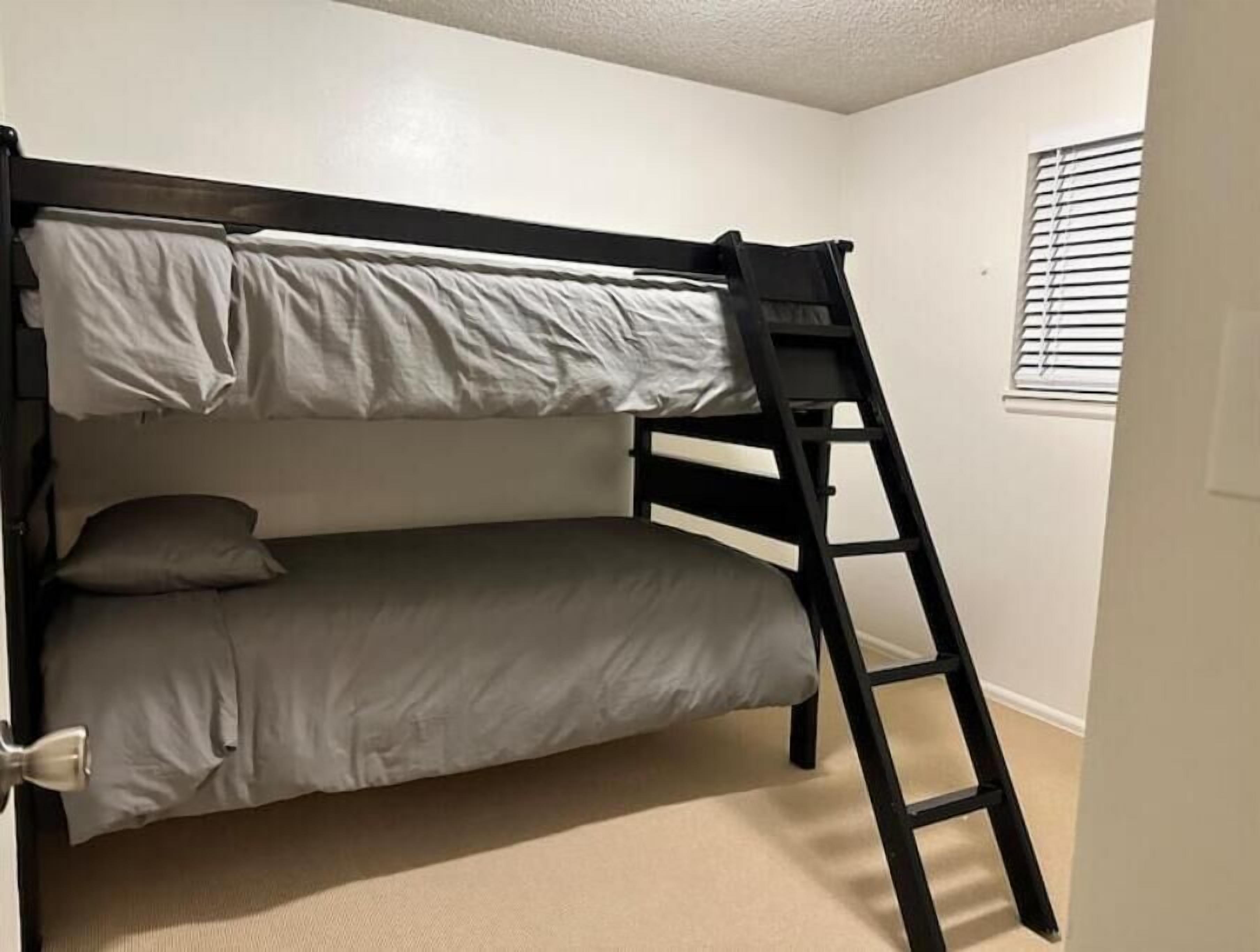 A bunk bed with a ladder, white walls, and a window with blinds.