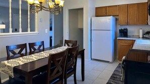 Un comedor con candelabro, sillas de madera, una mesa con mantel estampado y un refrigerador en la cocina contigua.