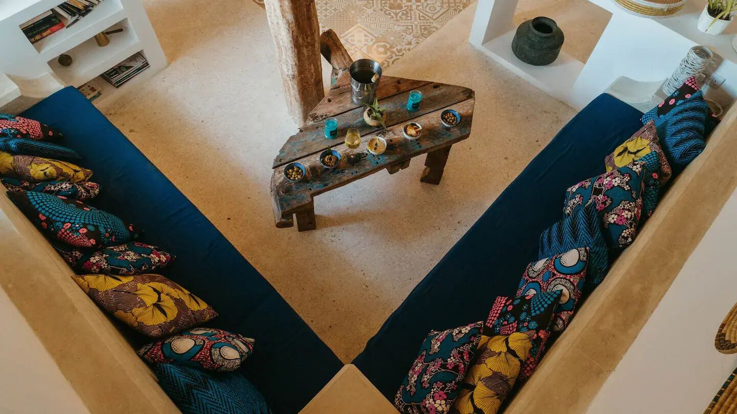 A cozy living area with a sofa set featuring vibrant patterned cushions, a rustic wooden coffee table with small bowls and a metal pitcher, and a white shelving unit with books and decorative items.