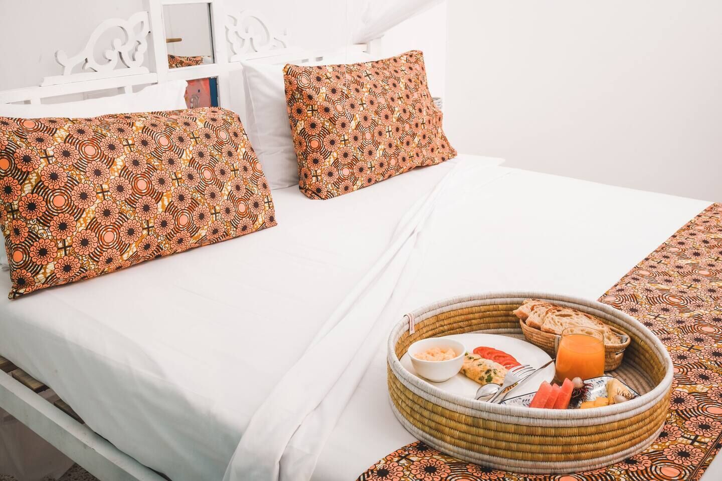 Cama bem arrumada com almofadas estampadas e um cesto de vime com itens de café da manhã sobre um lençol branco.