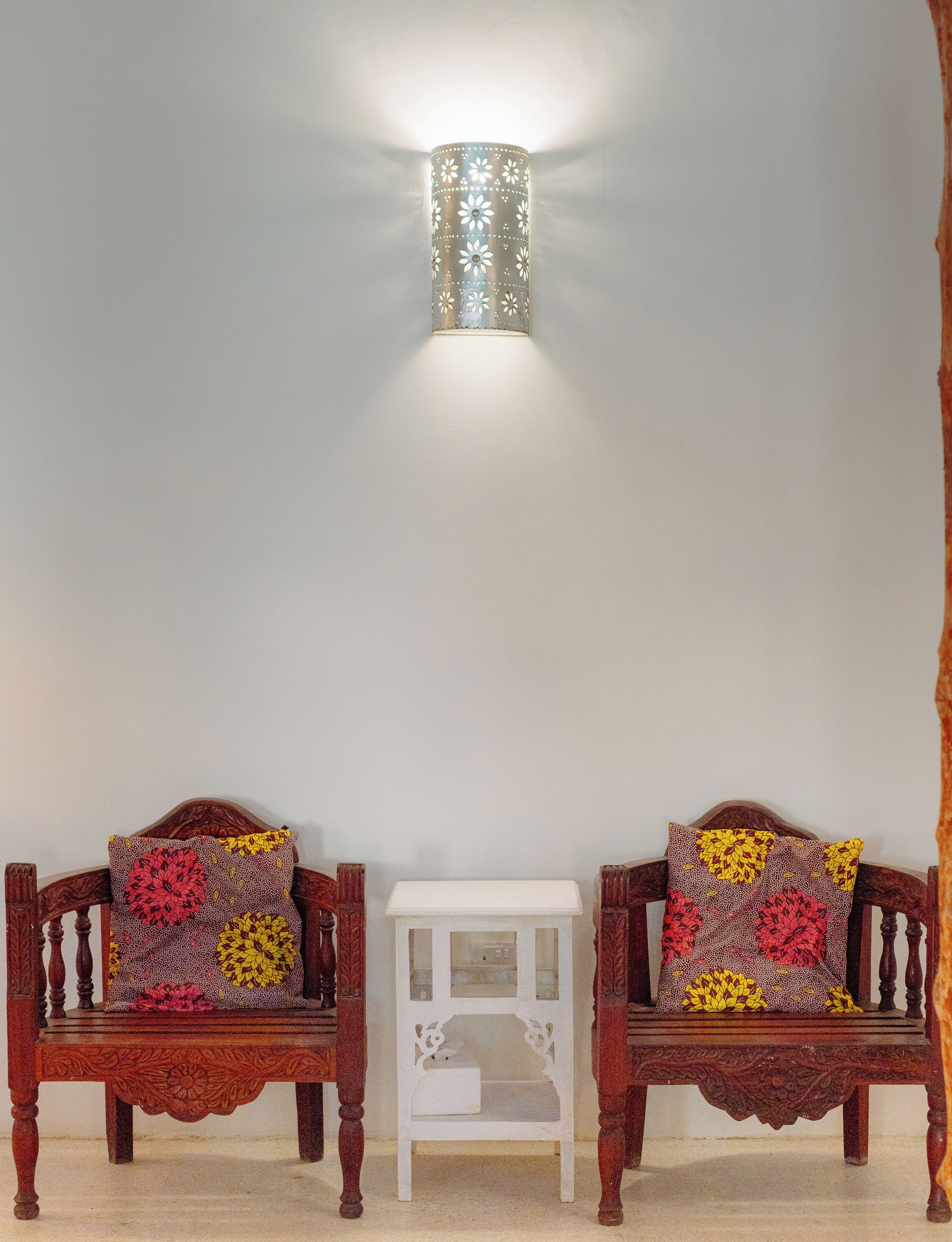 Two wooden chairs with floral cushions, a small white side table, and a wall-mounted light fixture.