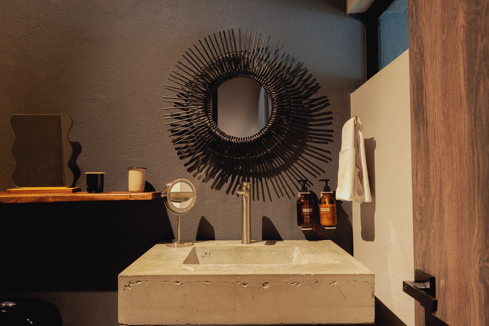 A bathroom with a central sink, a mirror with a sunburst frame, a towel hanging on the wall, and a shelf with toiletries.