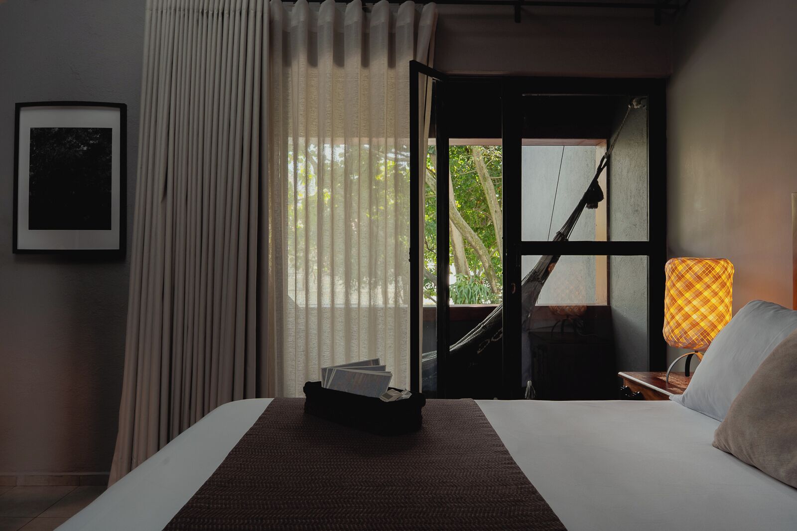 Habitación de hotel con una cama, una mesita de noche con lámpara, una ventana con cortinas y un cuadro en la pared.