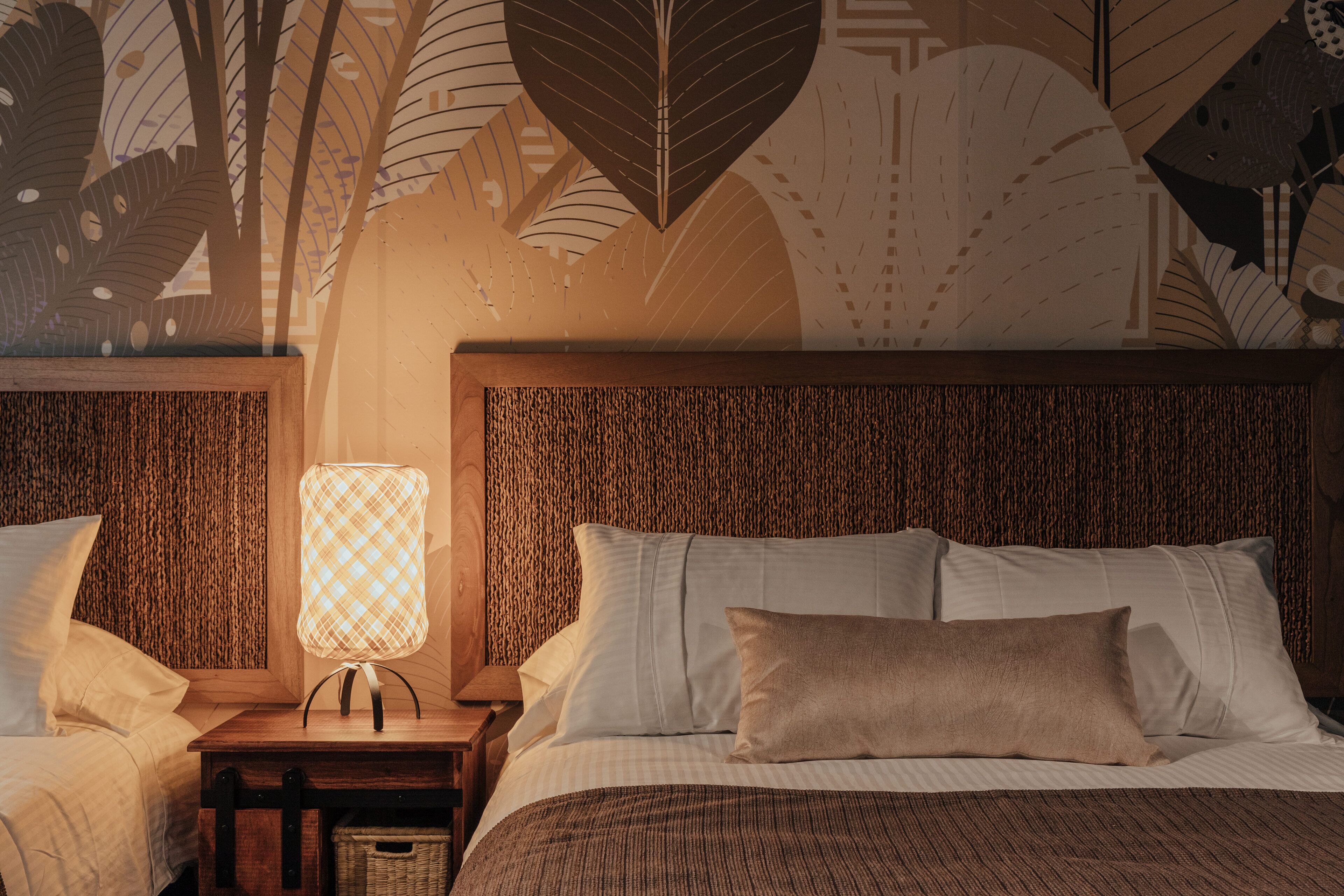 Una cama bien hecha con sábanas blancas y un almohadón marrón, una mesita de noche con una lámpara, y una pared con un diseño de hojas.