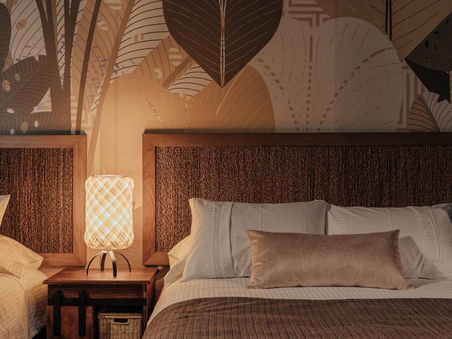Una cama bien hecha con sábanas blancas y un almohadón marrón, una mesita de noche con una lámpara, y una pared con un diseño de hojas.