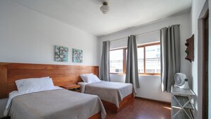 Ein Hotelzimmer mit zwei Betten, einem Holzkopfteil, einem kleinen Tisch mit Ventilator und einem Fenster mit Vorhängen.