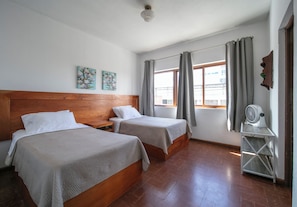 Habitación de hotel con dos camas, cabecero de madera, una mesita con un ventilador y una ventana con cortinas.