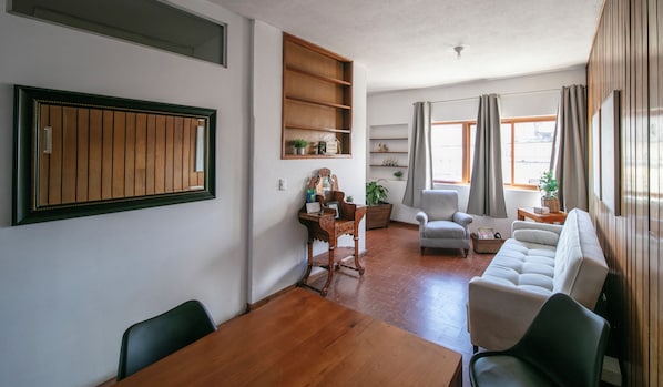 Ein Wohnzimmer mit einem Holzesstisch, einer Couch, einem Sessel, einem kleinen Beistelltisch und einem Regal mit Dekoration.