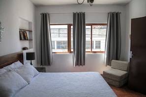 Ein Hotelzimmer mit einem Bett, einem Sessel, einem Nachttisch, einer Lampe und einem fensterbepflanzten Fenster.