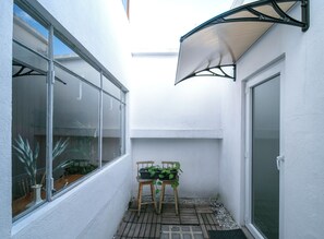 Un pequeño patio con una silla de madera y plantas en macetas, una pared blanca y una puerta corrediza de vidrio.