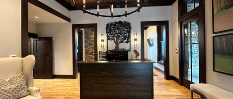A spacious room with a wooden floor, a central dark wooden island, a white sofa, a bench, and a large chandelier.