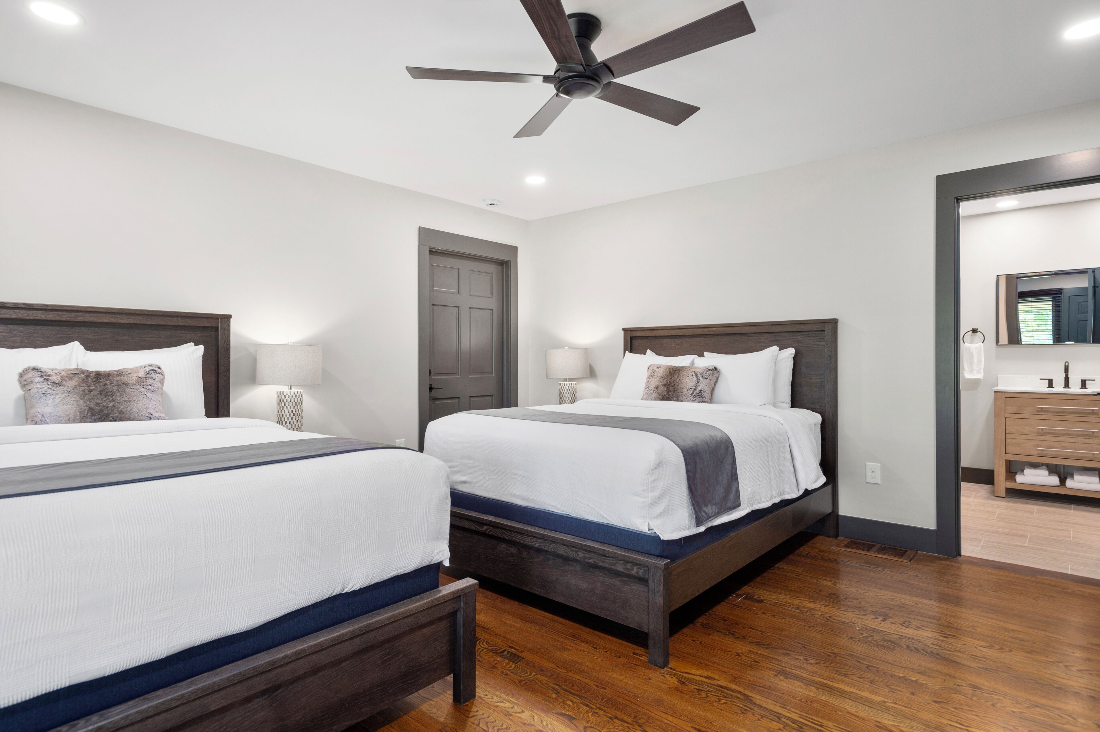 A hotel room with two beds, wooden floors, a ceiling fan, and a bathroom with a mirror and sink.