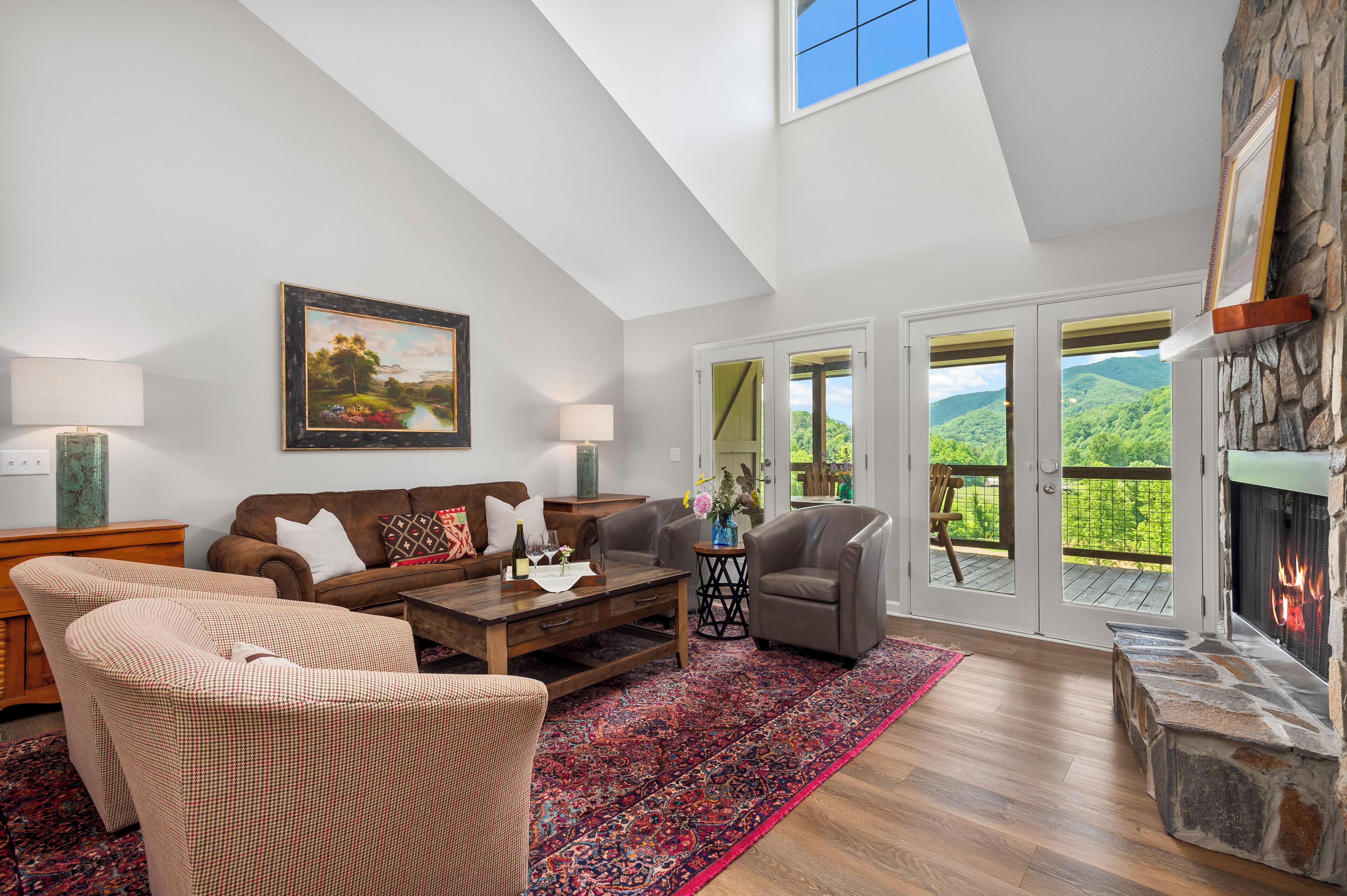 A spacious living room with a high ceiling, a fireplace, a large window with a view of greenery, and a cozy seating area.