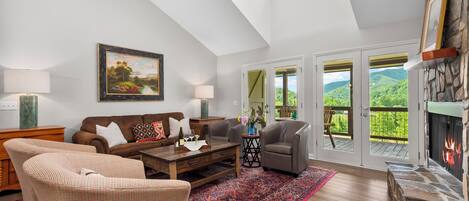 A spacious living room with a high ceiling, a fireplace, a large window with a view of greenery, and a cozy seating area.