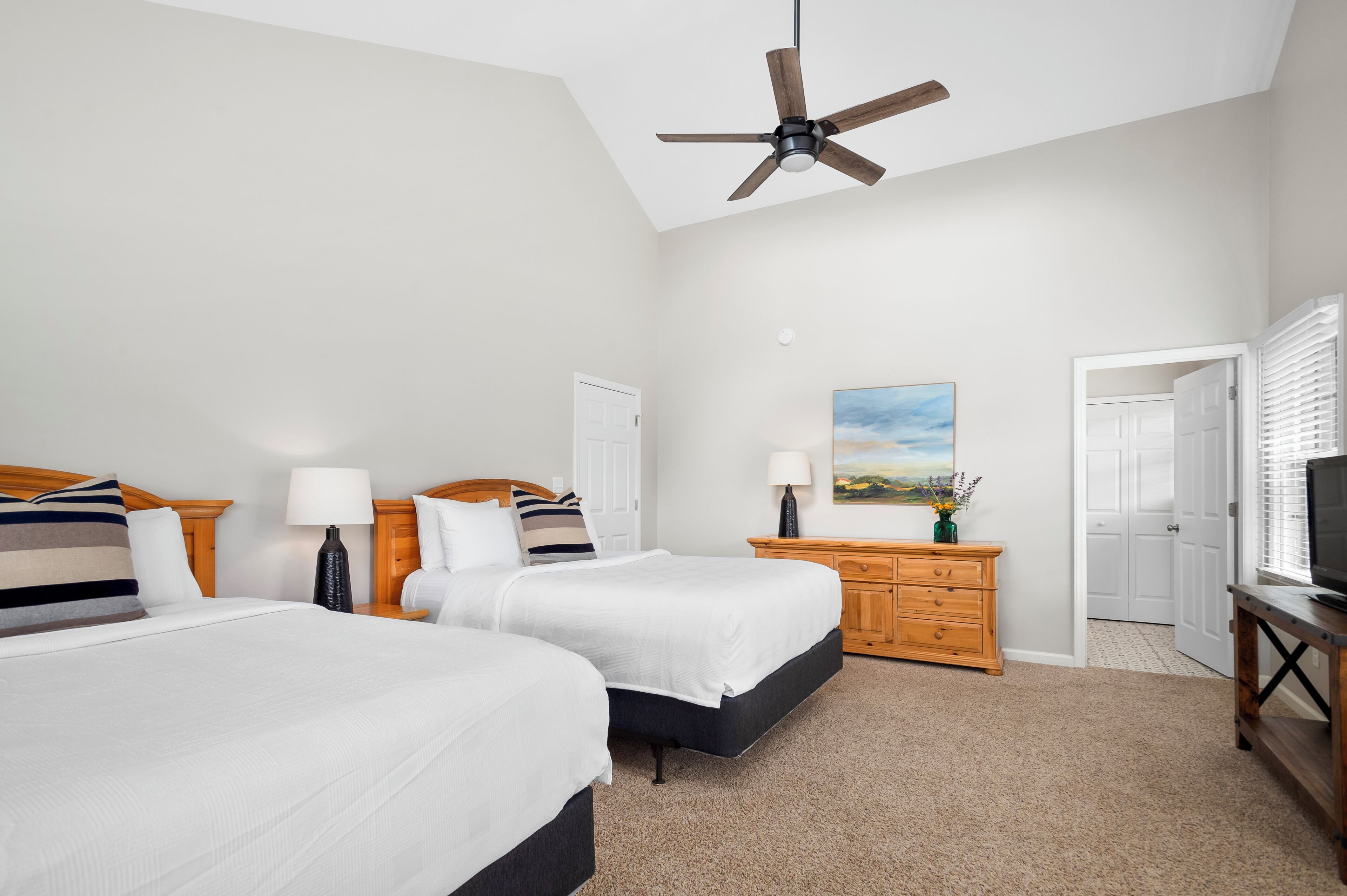 A hotel room with two beds, a ceiling fan, a painting, a TV, and a wooden dresser.