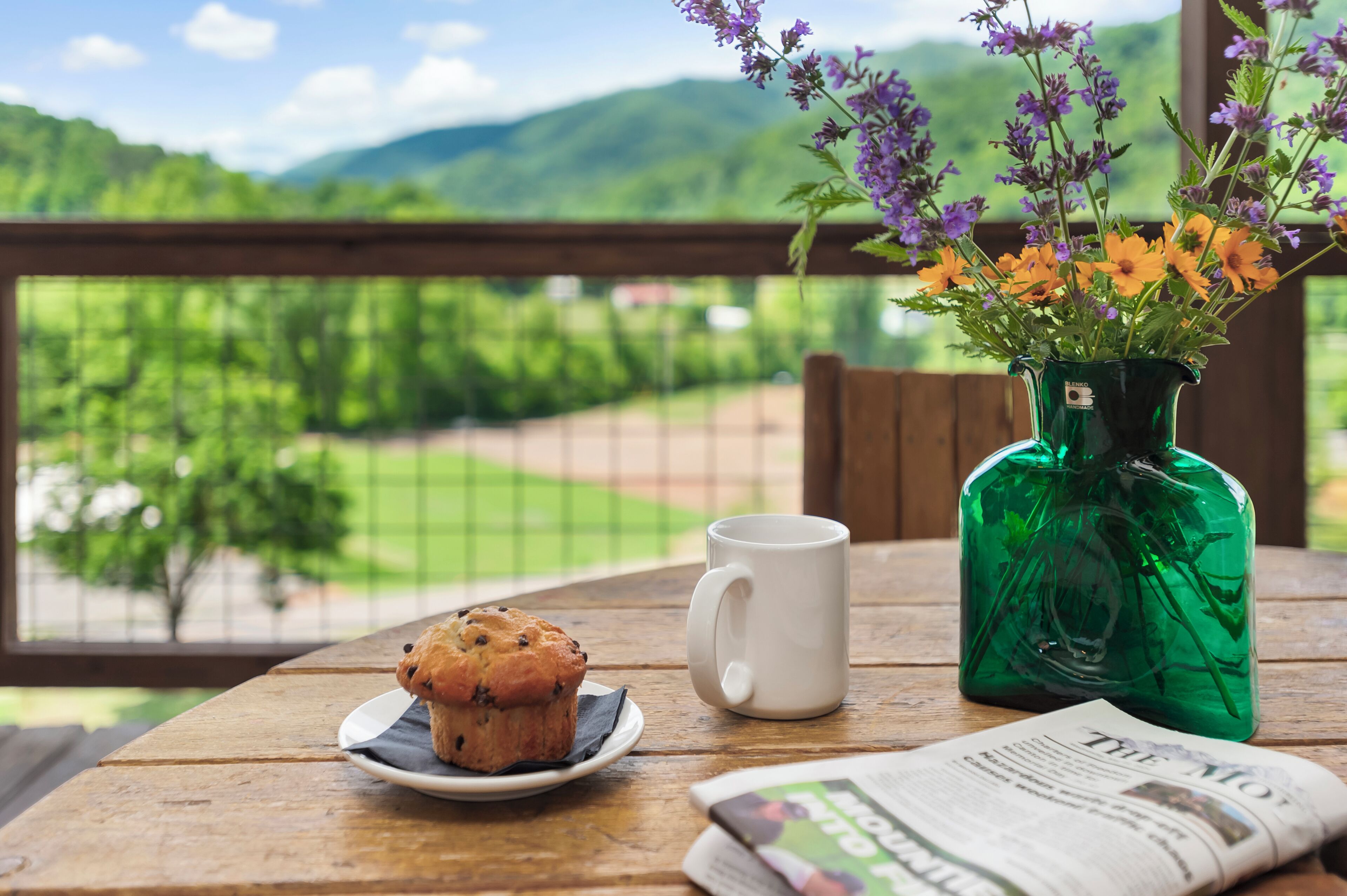 A wooden table with a muffin on a plate, a white mug, and a newspaper. A green vase with purple and orange flowers is also visible.