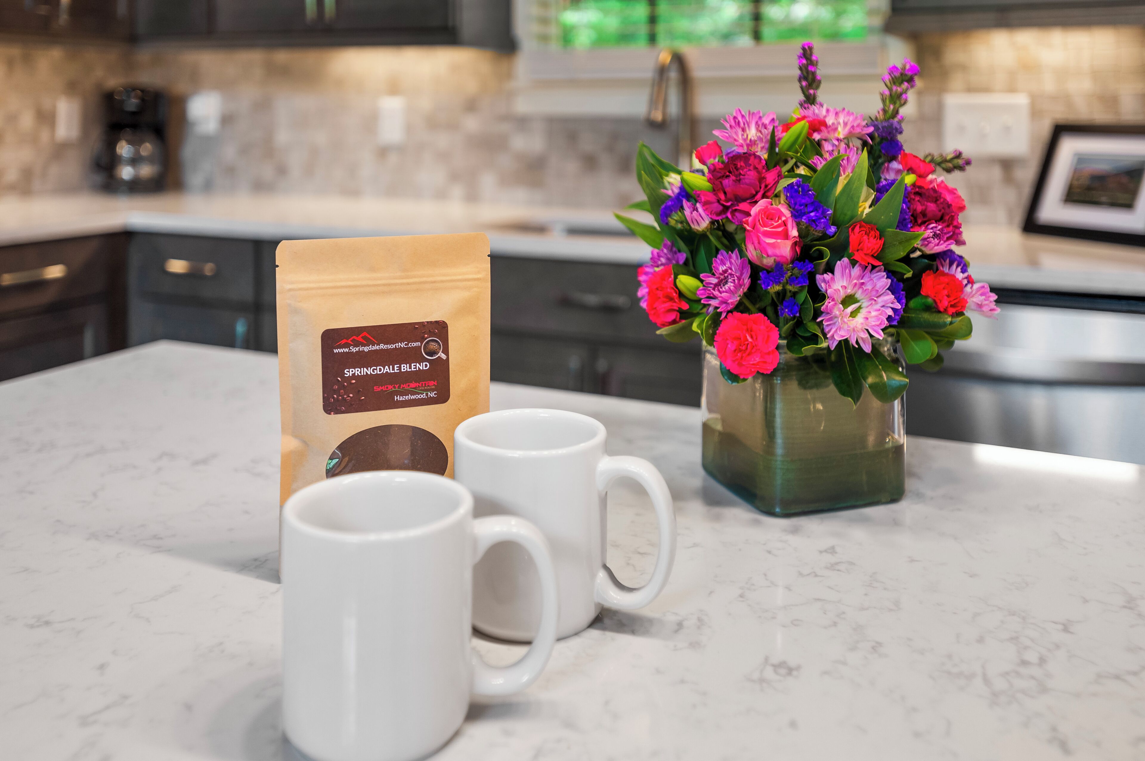 A kitchen counter with a coffee bag, two white mugs, and a vase of colorful flowers.