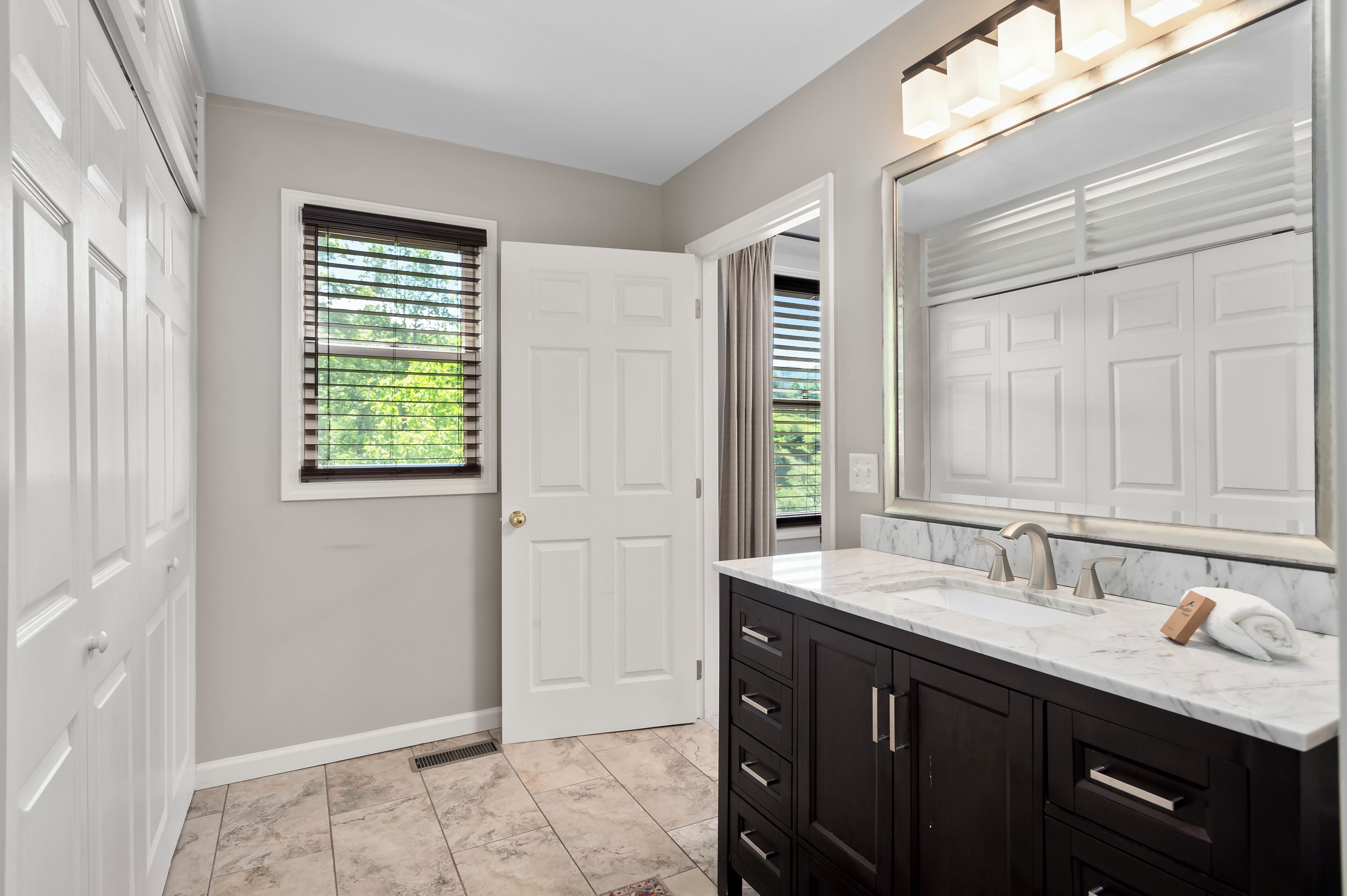 A bathroom with a white door, a window with blinds, a sink with a marble countertop, and dark cabinets.