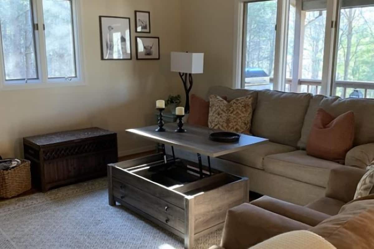 A living room with a sofa, a coffee table, a side table with candles, and a lamp. There are framed pictures on the wall and a window with a view of trees.
