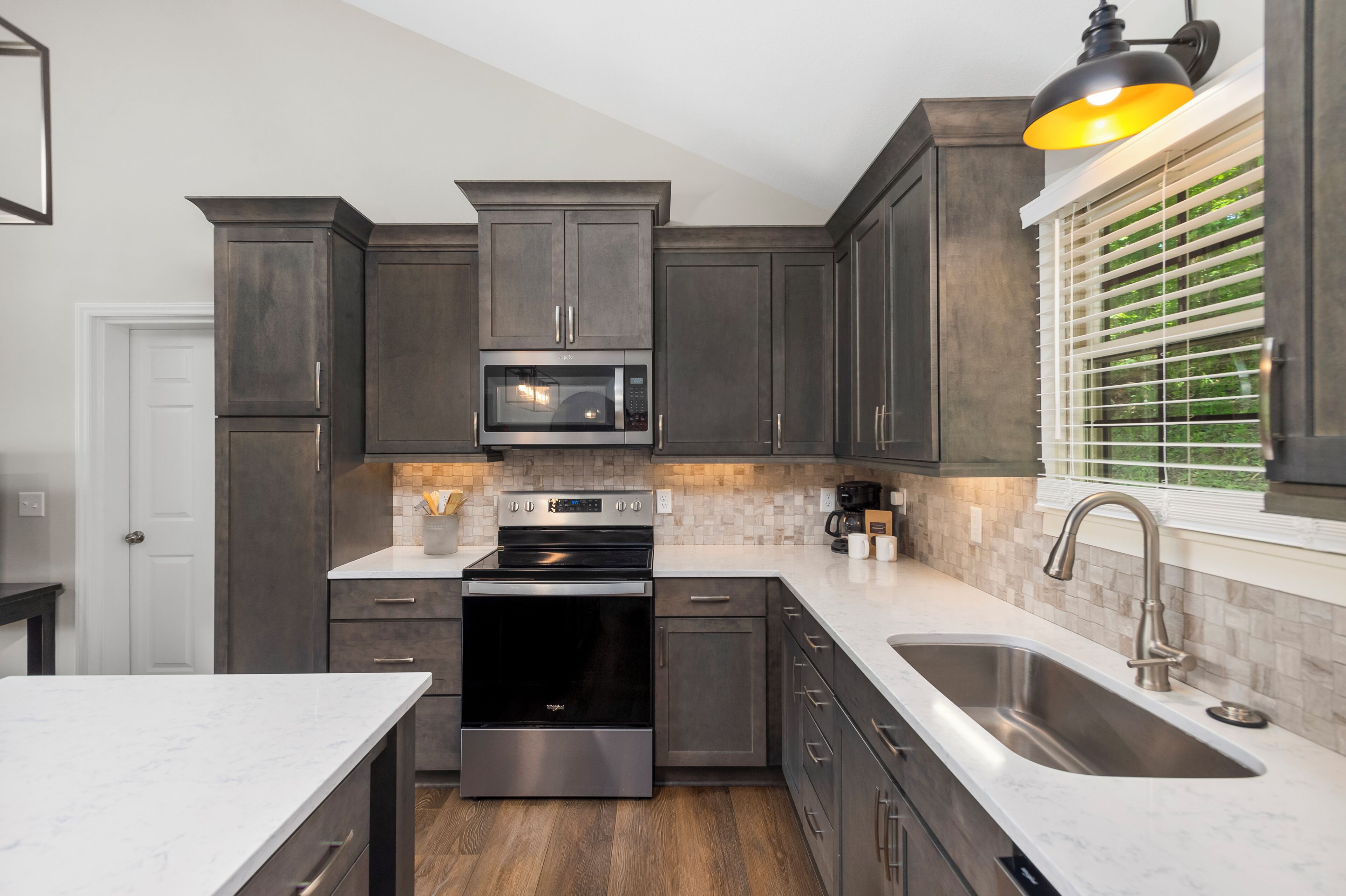 A modern kitchen with dark cabinetry, stainless steel appliances, and a large window with blinds.