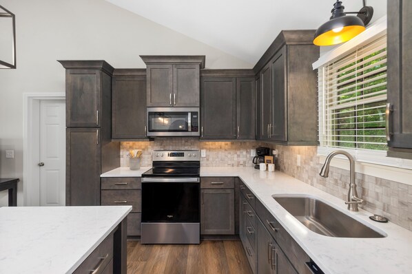 A modern kitchen with dark cabinetry, stainless steel appliances, and a large window with blinds.