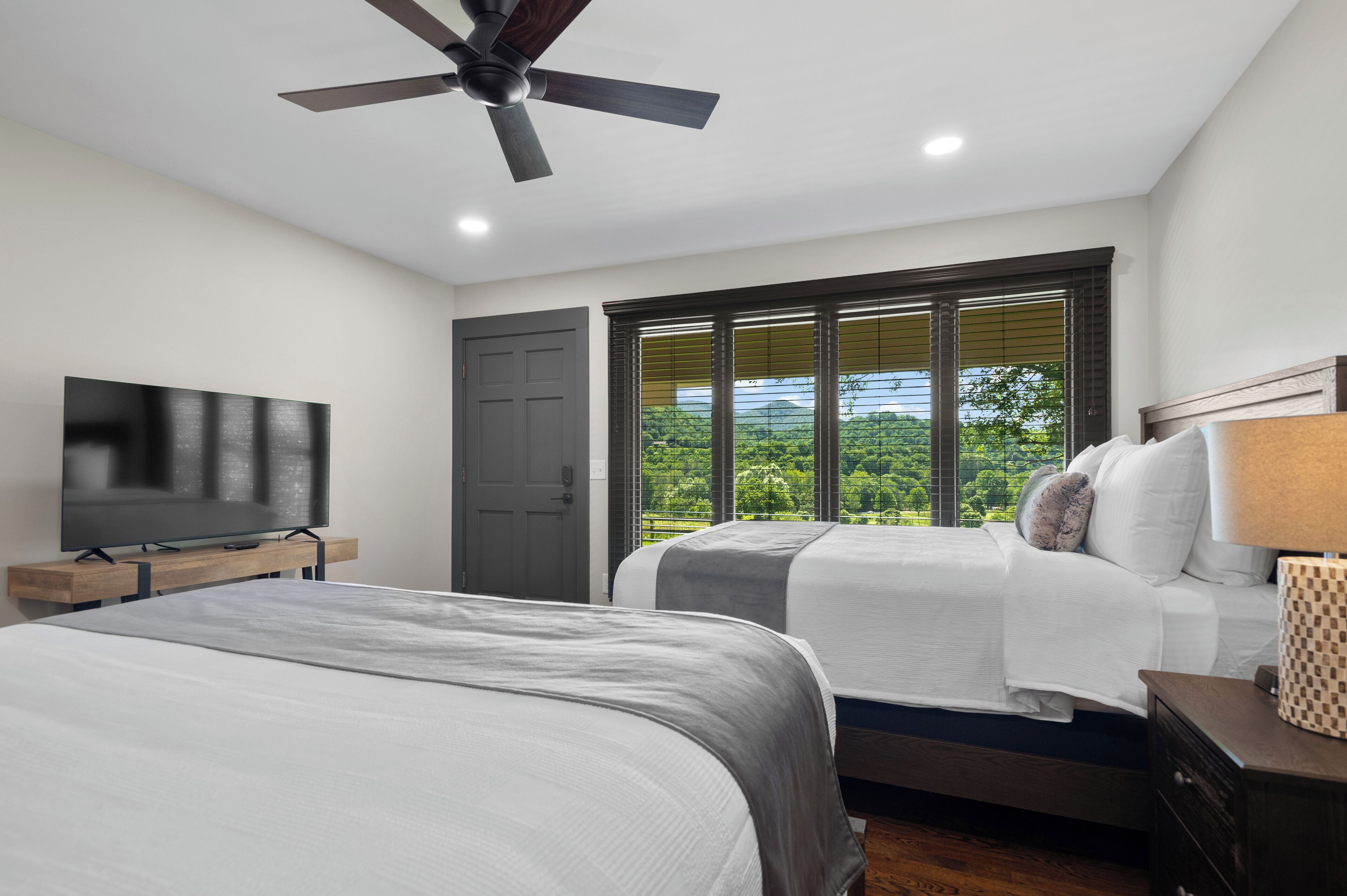 A bedroom with two beds, a ceiling fan, a TV, and a window with a view of greenery.